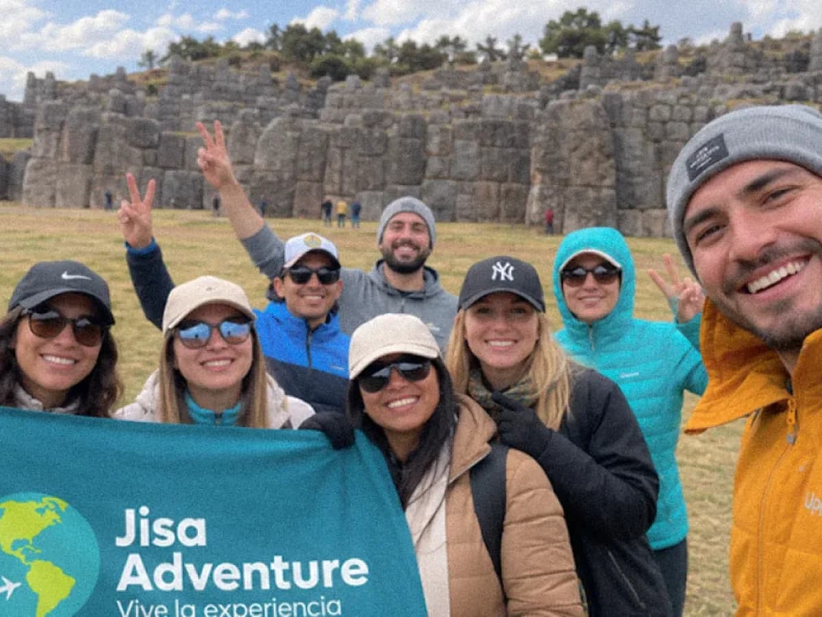 Turistas disfrutando del City Tour Cusco en Sacsayhuamán, posando con la bandera de Jisa Adventure frente a los muros incas.